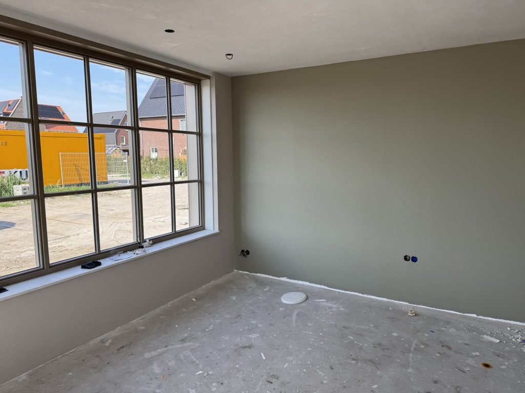 Nieuwbouwkamer met een egaal geverfde grijsgroene wand en een breed raam met roedeverdeling