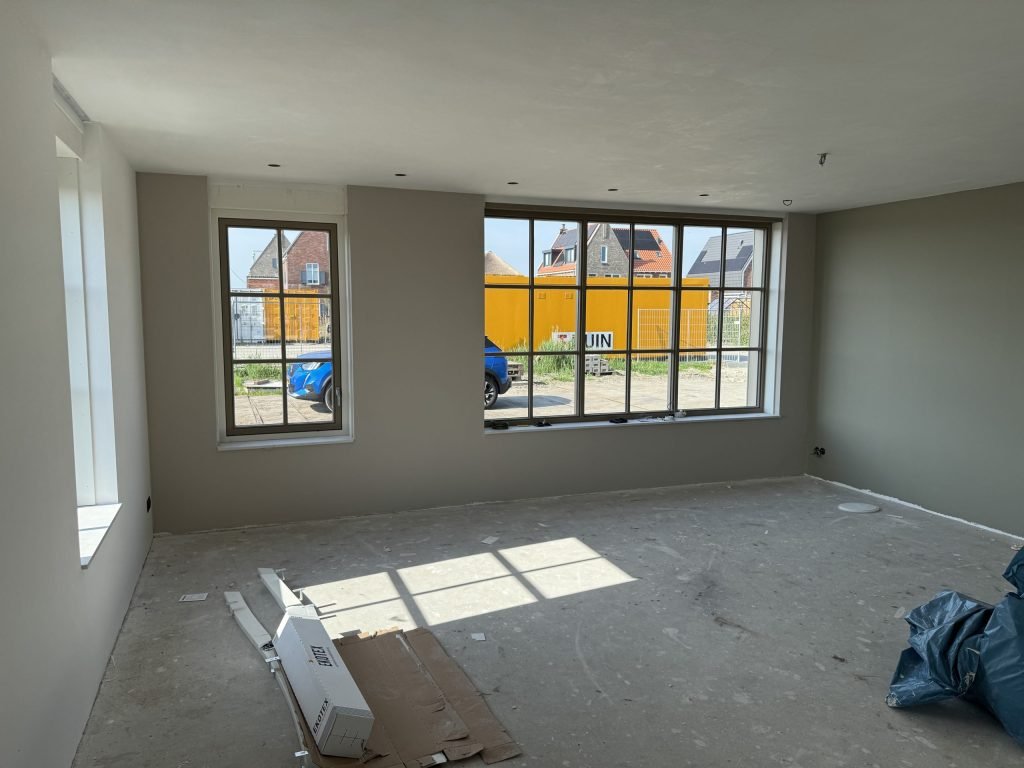 Opgeleverde nieuwbouwwoonkamer met grijsgroene wanden, kale betonvloer en ramen met roedeverdeling
