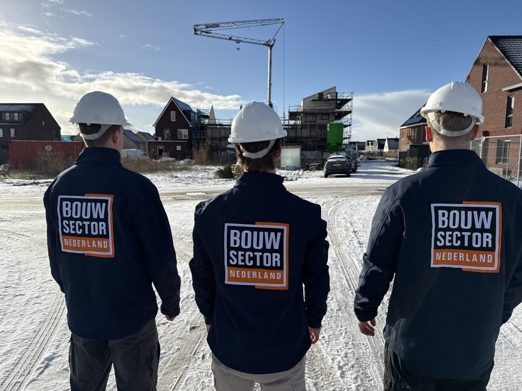 Drie vakmensen van Bouwsector Nederland met witte helm en logo op de rug in een besneeuwde straat met nieuwbouwhuizen en een bouwkraan