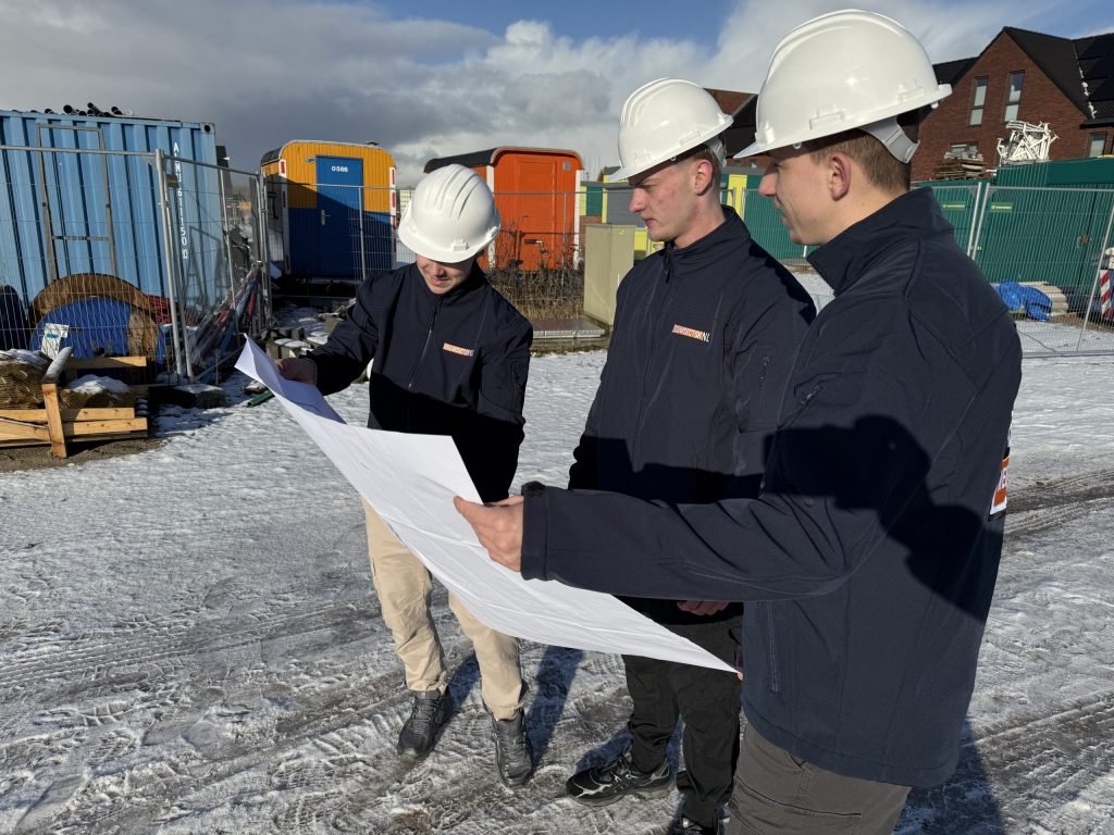 Drie vakmensen van Bouwsector Nederland met witte helm bekijken samen een plattegrond op een besneeuwde bouwplaats